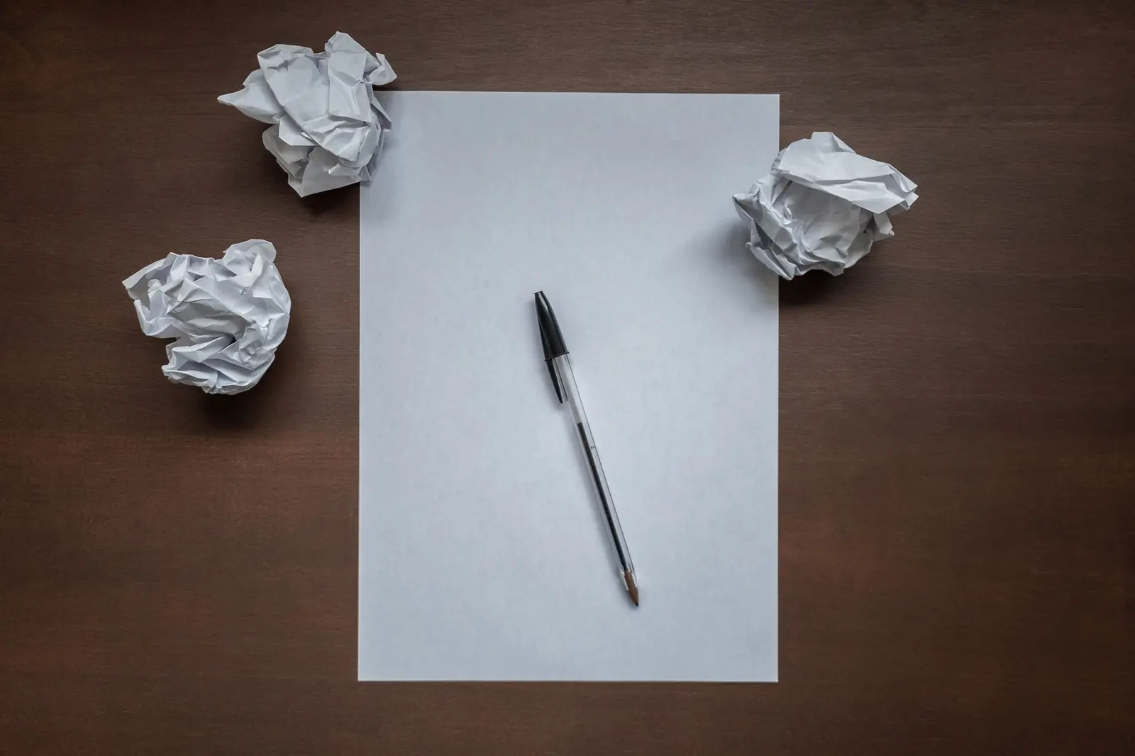 A blank sheet of paper and a pen on a dark desk, with three crumpled drafts beside it